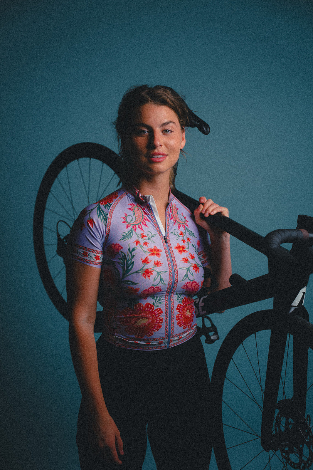 Woman wearing a colorful cycling jersey holding a bicycle against a blue background
